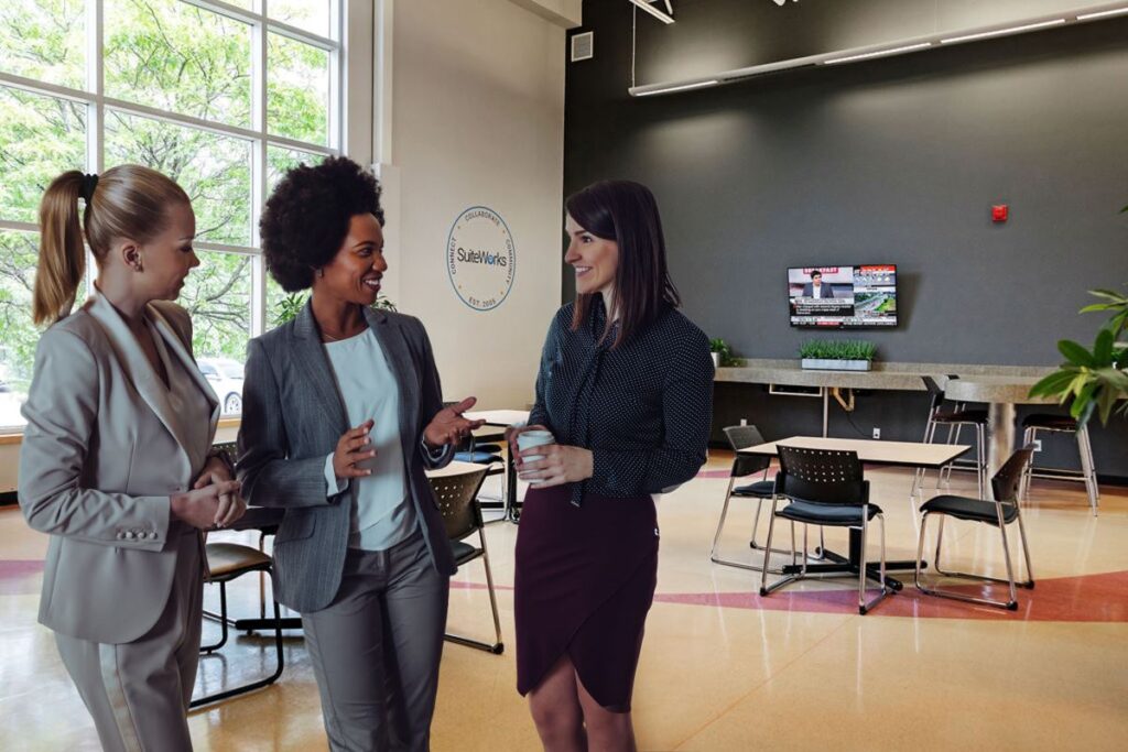 three women are standing in a well-lit office area at SuiteWorks, with a large window providing a view and ample natural light.